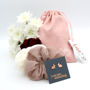 Pink scrunchie, rose gold bunny earrings, and flowers with a 'Sister & Co' drawstring bag on a white background.