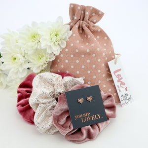 Three scrunchies in pink, white, and rose gold with a polka dot bag and decorative card on a white background.