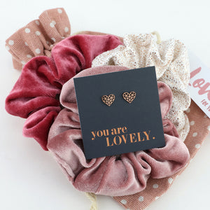 Pink scrunchies with heart-shaped earrings and a 'you are LOVELY.' card on a white background