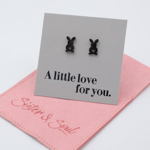 Black bunny-shaped earrings on a gray card with 'A little love for you' text, placed on a pink cloth with 'Sister & Swind' branding.