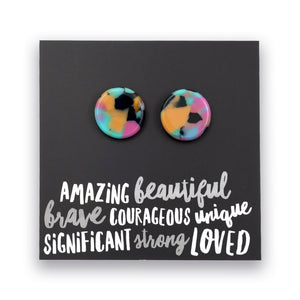 A pair of round stud earrings with a colorful marbled pattern in pink, yellow, blue, and black, displayed on a black card with white text that reads 'AMAZING beautiful brave COURAGEOUS unique SIGNIFICANT strong LOVED'.