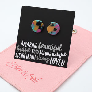 A pair of round stud earrings with a colorful marbled pattern in pink, blue, yellow, and black, displayed on a black card with inspirational text. The card is resting on a pink fabric pouch embroidered with 'Sister & Soul'.