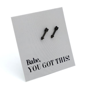 A pair of black arrow-shaped stud earrings displayed on a textured light gray square card that reads 'Babe, YOU GOT THIS!' in black text, photographed on a white background.