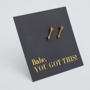 A pair of small gold arrow-shaped stud earrings displayed on a dark charcoal textured backing card with gold text that reads 'Babe, YOU GOT THIS!' on a light gray background.