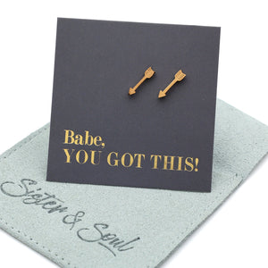 Two gold arrow-shaped stud earrings displayed on a dark charcoal card that reads 'Babe, YOU GOT THIS!' in gold lettering, resting on a light sage green jewelry pouch with 'Sister & Soul' written on it.