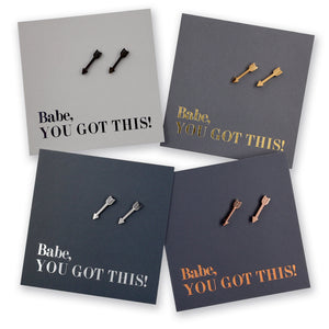 Four square product cards arranged in a 2x2 grid, each showing a pair of small arrow-shaped stud earrings on a dark card with the phrase 'Babe, YOU GOT THIS!' in metallic lettering. The earrings and cards come in black, gold, silver, and rose gold colorways.