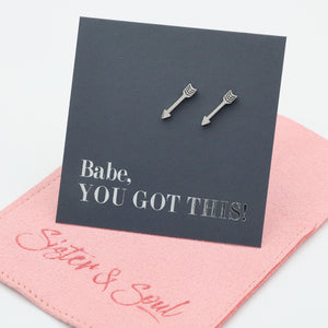 A pair of silver arrow stud earrings displayed on a dark gray card that reads 'Babe, YOU GOT THIS!' with a pink fabric pouch underneath.