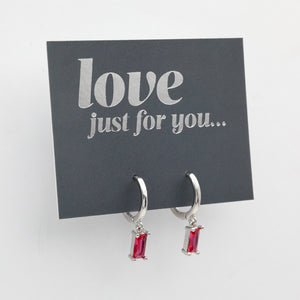 A pair of silver huggie hoop earrings with rectangular red gemstones, displayed on a dark gray card that reads 'love just for you.