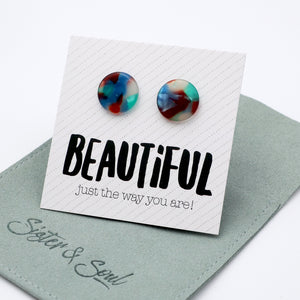 A pair of round stud earrings with a marbled swirl pattern of blue, red, and teal, displayed on a white branded card that reads 'BEAUTIFUL just the way you are!' resting on a sage green pouch from Sister & Soul.