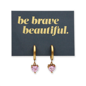 A pair of gold hoop earrings with dangling pink heart-shaped crystals, displayed on a dark gray card that reads 'be brave beautiful.' in gold lettering, against a white background.