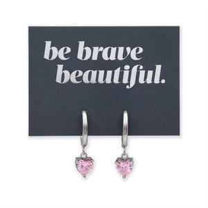 A pair of silver-toned drop earrings with small heart-shaped pink gemstones, displayed on a dark charcoal card that reads 'be brave beautiful.' in white text, against a plain white background.