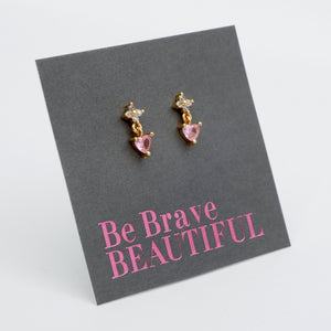 A pair of gold dangle earrings with pink heart-shaped crystals and small clear stones, displayed on a dark gray card that reads 'Be Brave BEAUTIFUL' in pink text, on a white background.