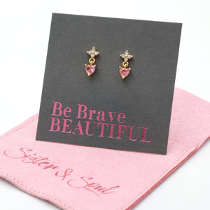 Gold-tone dangle earrings with a small clear crystal star and a pink heart-shaped crystal drop, displayed on a dark gray card that reads 'Be Brave BEAUTIFUL' above a pink branded jewelry pouch.