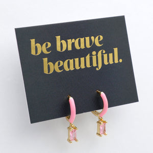 Pink hoop earrings with gold accents on a black card with 'be brave beautiful' text.