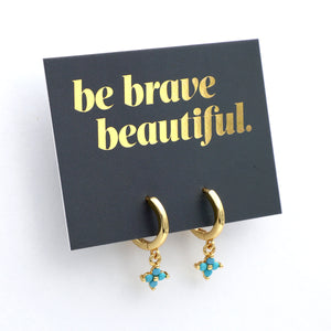 Gold earrings with turquoise charms on a black card with 'be brave beautiful' text.