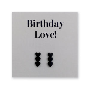 A pair of black heart-shaped stud earrings displayed on a dark charcoal gray gift card that reads 'Birthday Love!' in black lettering.
