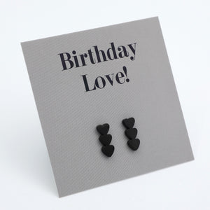 Black heart stud earrings displayed on a textured gray card that reads 'Birthday Love!' in black serif text, photographed on a clean white background.