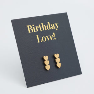 A pair of small gold heart stud earrings displayed on a dark charcoal card that reads 'Birthday Love!' in gold lettering, photographed on a light gray background.