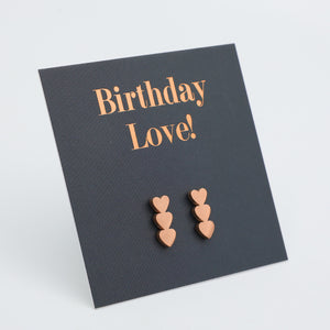 A pair of rose gold heart stud earrings displayed on a dark charcoal gray card that reads 'Birthday Love!' in rose gold lettering, set against a clean white background.
