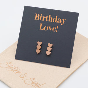 Three-pair heart stud earrings in a rose gold tone, displayed on a black square card that reads 'Birthday Love!' in rose-gold lettering, resting on a beige pouch with the script 'Sister & Soul'.
