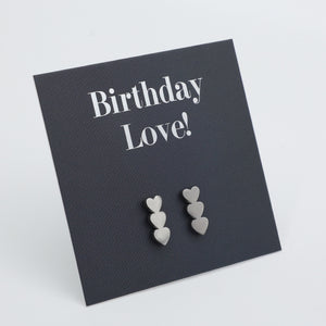 Three pairs of small silver heart stud earrings displayed on a dark charcoal square card that reads 'Birthday Love!' in white text, photographed on a light gray background.