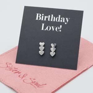 A pair of silver heart drop stud earrings displayed on a black card that reads 'Birthday Love!' with a pink fabric pouch underneath on a light gray background.