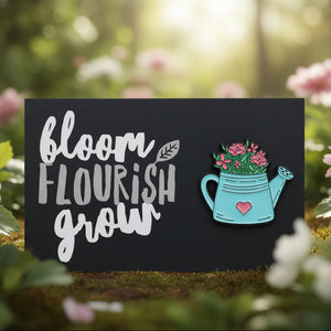 A rectangular black card with the words “bloom flourish grow” in white lettering, with a turquoise watering can with pink flowers and a small pink heart.