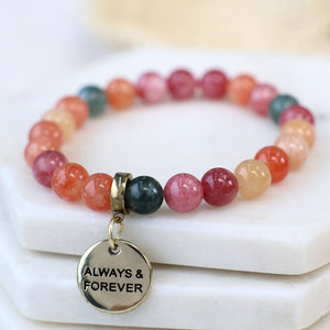A beaded stretch bracelet with round gemstone-like beads in pink, orange, red, and dark green, featuring a gold-tone charm engraved with 'ALWAYS & FOREVER', displayed on stacked white stone blocks.