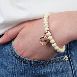 White beaded bracelet with a rose gold charm labeled 'Dream' on a wrist.