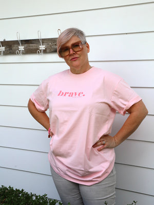 A woman with short gray hair and sunglasses wearing a light pink t-shirt with the word 'brave.' printed in darker pink across the chest, standing against a white siding wall.