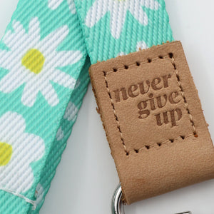 Green floral-patterned belt with a brown leather tag reading 'never give up' on a white background