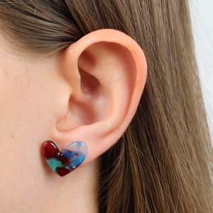 A close-up of a person's ear wearing a small heart-shaped stud earring with a marbled swirl pattern of blue, red, and teal.