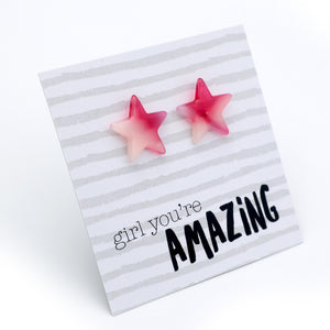 A pair of pink gradient star-shaped stud earrings displayed on a white and gray striped card that reads 'girl you're AMAZING' on a clean white background.