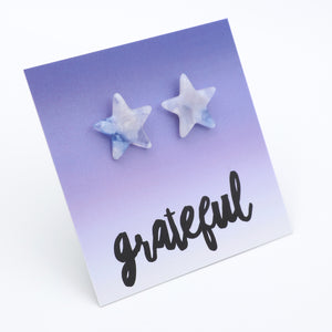 A pair of star-shaped stud earrings with a light blue and white marbled pattern, displayed on a purple-to-white gradient card that features the word 'grateful' in black script.