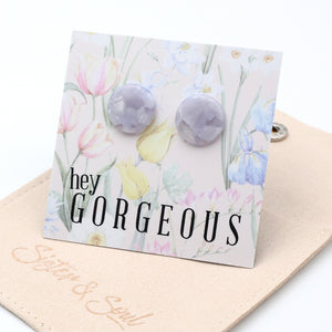 A pair of round lavender-gray stud earrings displayed on a floral backing card that reads 'hey GORGEOUS.' The card is placed on a beige fabric surface with the brand name 'System & Soul'