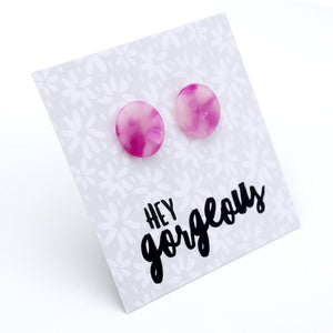 A pair of round stud earrings with a pink and purple marbled pattern, displayed on a white card with a floral pattern and the text 'HEY gorgeous' in black script.