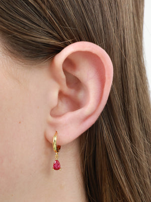 A close-up of a person's ear wearing a small gold huggie hoop earring with a dangling teardrop-shaped red gemstone.