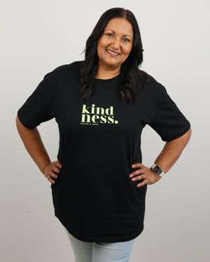 A smiling woman with dark hair stands with hands on hips wearing a black short-sleeve t-shirt with 'kindness.' printed in yellow-green text on the chest, paired with light blue jeans, against a plain light gray background.