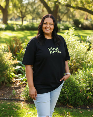 A woman wearing a black graphic t-shirt with the words 'kindness.' in yellow-green text, paired with light blue jeans, standing against a plain light gray background.