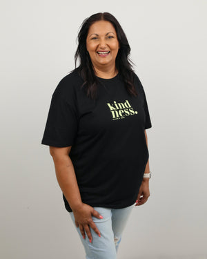 A smiling woman with dark hair stands against a plain light gray wall wearing a black short-sleeve t-shirt with 'kindness.' printed in yellow-green text on the chest, paired with light blue jeans.