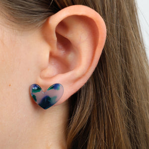 A close-up of a person's ear wearing a small heart-shaped stud earring with a marbled pattern of blue, green, and translucent pink.