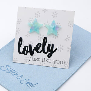Two iridescent star-shaped stud earrings displayed on a white card that reads 'lovely just like you!' with a blue fabric pouch underneath. The earrings have a pastel blue and mint green color with subtle purple iridescence.