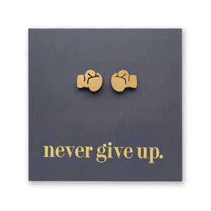 A pair of small gold stud earrings shaped like boxing gloves, displayed on a dark gray square backing card with the phrase "never give up." in gold lowercase text.