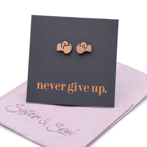 A pair of small rose gold stud earrings shaped like boxing gloves, displayed on a dark charcoal card that reads 'never give up.' in rose gold lettering, resting on a purple fabric pouch with the brand name 'Sister & Soul' visible.
