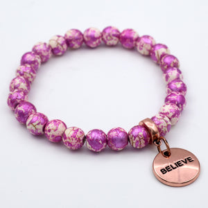 Purple and white beaded bracelet with a rose gold 'BELIEVE' charm on a light gray background