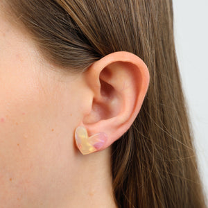 A close-up of a person's ear wearing a small translucent iridescent clip-on earring in a pale yellow-pink color.