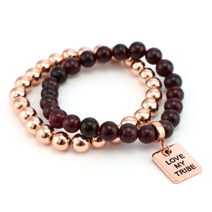 Two bracelets stacked together, one with smooth red garnet beads and the other with rose gold-colored metal beads, ending with a charm that reads 'LOVE MY TRIBE'.