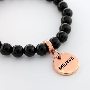 Black beaded bracelet with a rose gold charm labeled 'BELIEVE' on a white background