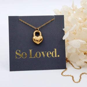 Gold heart-shaped lock necklace on a card with 'So Loved' text, surrounded by white flowers.