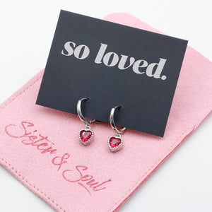 Pink heart shaped huggies dangle earrings presented on a grey card with silver 'so loved' writing sitting on a pink 'Sister & Soul' pouch.